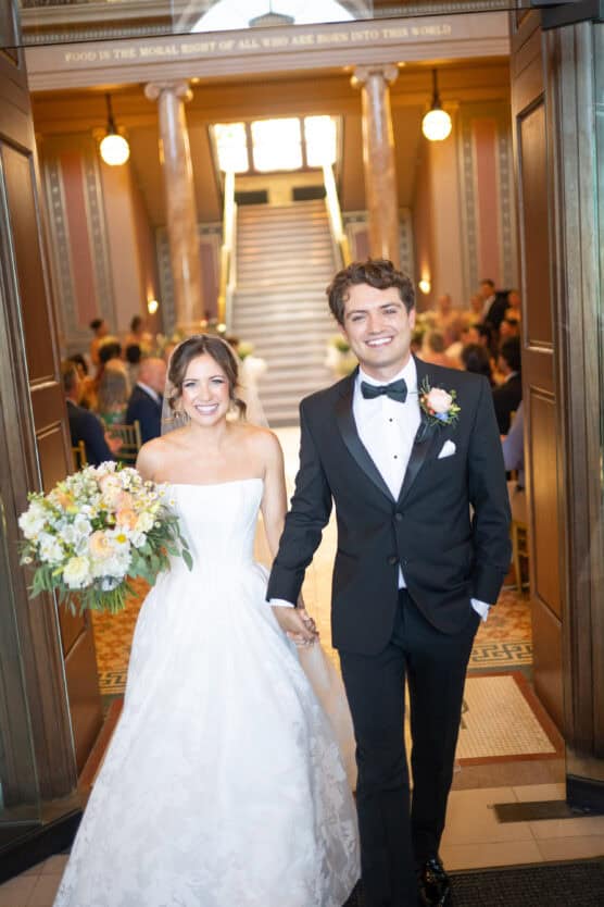 A smiling bride in a white strapless gown and a groom in a black tuxedo walk hand in hand down an aisle, with guests seated in the background inside a grand, elegant hall. by Justin Salem Meyer