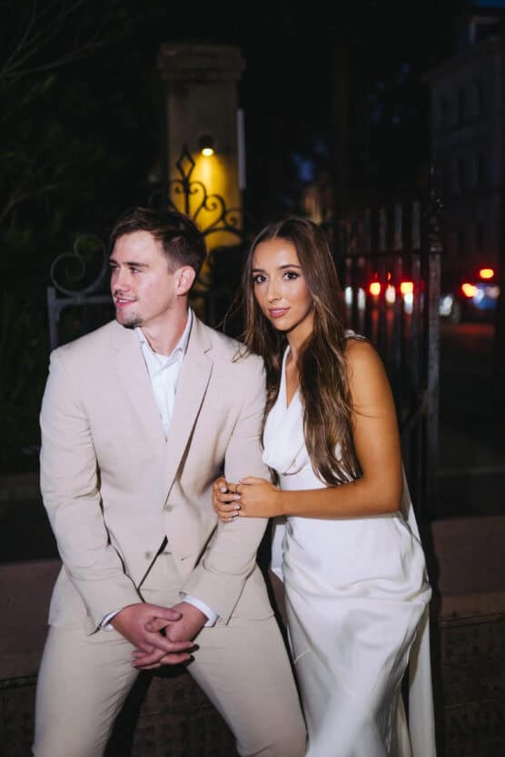 A man in a beige suit sits beside a woman in a white dress; she stands, holding his arm, and both face the camera at night with blurred streetlights in the background. by Justin Salem Meyer