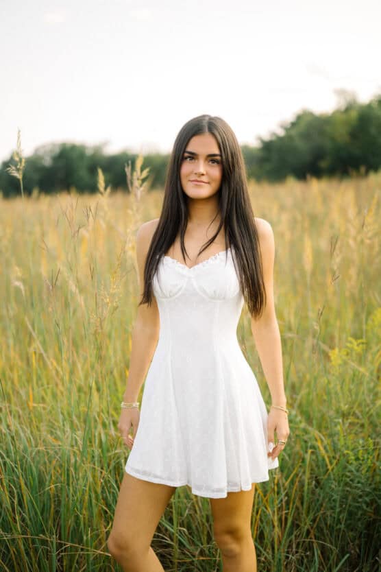 A young woman with long dark hair wearing a sleeveless white dress stands in a grassy field with tall green and yellow plants, trees blurred in the background, and soft natural light. by Justin Salem Meyer