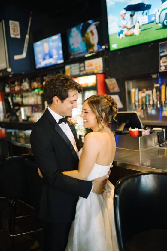 A bride and groom in wedding attire smile and embrace inside a sports bar, with TV screens showing sports above them and bar stools and counters in the background. by Justin Salem Meyer