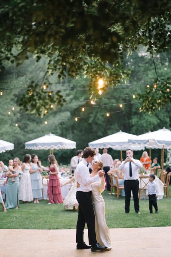 A couple dances together outdoors under string lights, surrounded by guests in formal attire. Tables with umbrellas and trees are in the background, creating a festive, romantic atmosphere. by Justin Salem Meyer