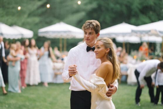 A young couple, dressed formally, dances outdoors on grass. The woman wears an off-the-shoulder white dress, and the man wears a white shirt with a black bow tie. Guests and white canopies are blurred in the background. by Justin Salem Meyer