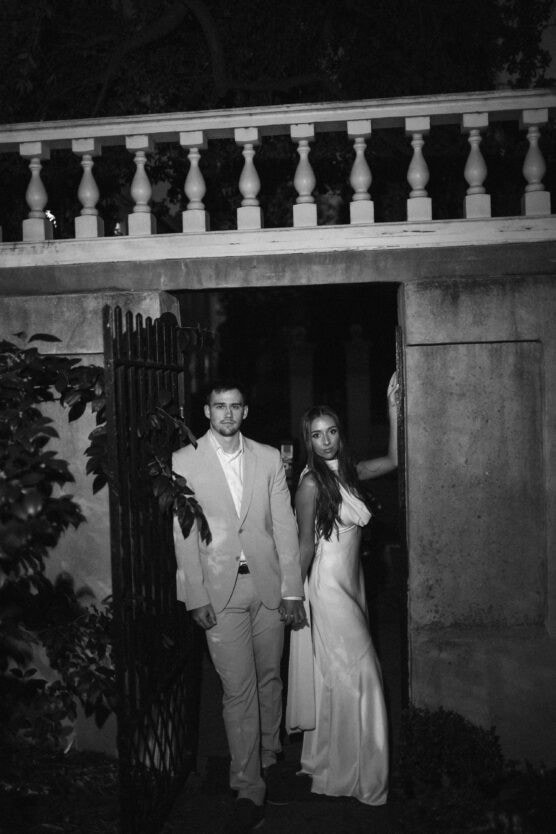 A man in a light suit and a woman in a satin dress stand holding hands in a doorway at night, illuminated by soft light, with a wrought iron gate and stone wall behind them. The scene is in black and white. by Justin Salem Meyer
