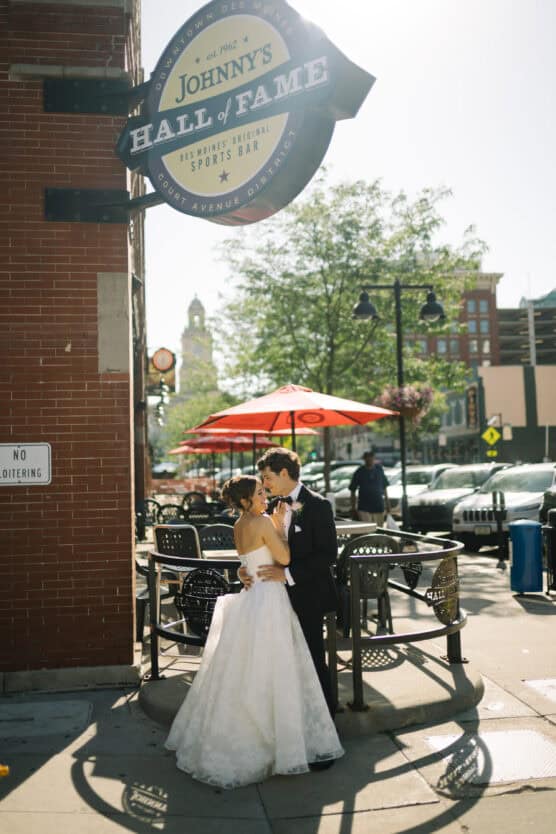 A bride and groom embrace and smile at each other on a sunny sidewalk outside Johnny’s Hall of Fame sports bar, with outdoor tables, red umbrellas, and city buildings in the background. by Justin Salem Meyer