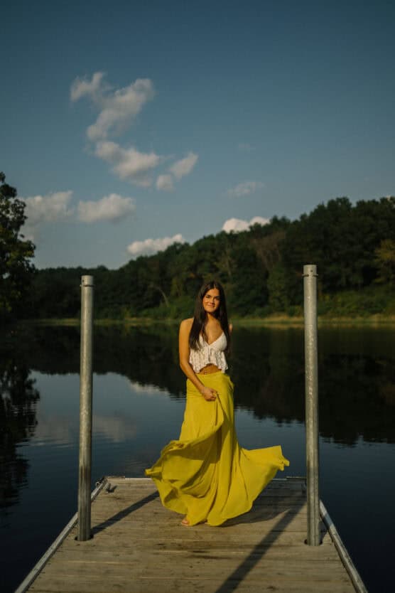 A woman in a white top and flowing yellow skirt stands barefoot on a wooden dock by a calm lake, with green trees and a blue sky with clouds in the background. by Justin Salem Meyer