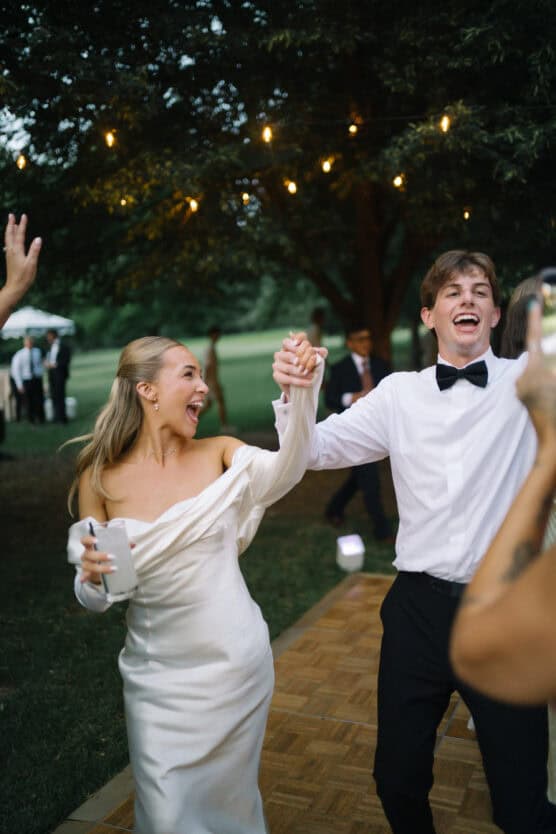 A joyful bride in an off-the-shoulder white dress and a groom in a white shirt and black bow tie hold hands and smile while dancing outdoors under string lights. by Justin Salem Meyer