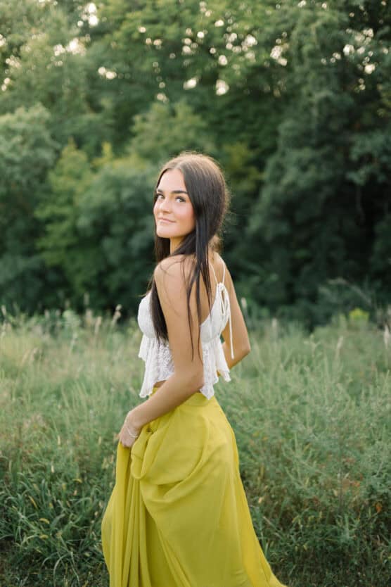 A young woman with long dark hair stands in a green field, wearing a white sleeveless top and a flowing yellow skirt, looking back over her shoulder. Lush trees fill the background. by Justin Salem Meyer