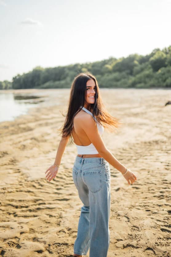 A young woman with long brown hair, wearing a white tank top and blue jeans, smiles and looks over her shoulder while walking on a sandy beach with trees in the background. by Justin Salem Meyer