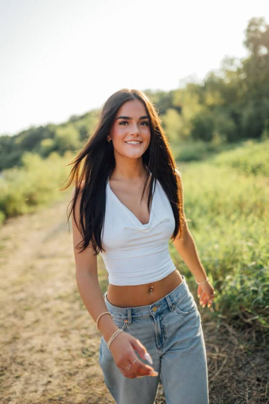 A young woman with long dark hair, wearing a white sleeveless top and blue jeans, smiles while walking outdoors on a sunny day with green trees and grass in the background. by Justin Salem Meyer