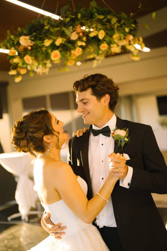 A bride and groom in formal attire share a joyful dance at their wedding reception, smiling at each other under a floral chandelier. by Justin Salem Meyer