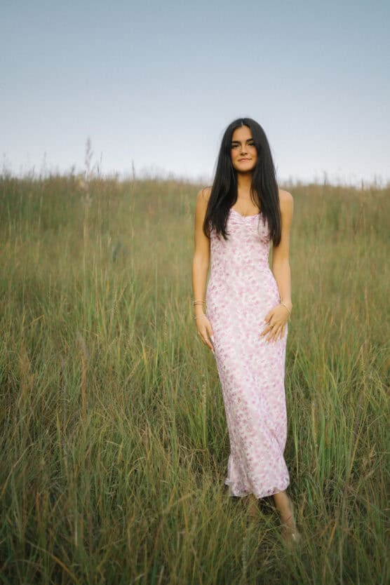 A woman with long dark hair wearing a light pink floral dress stands in a field of tall grass under a clear sky. by Justin Salem Meyer