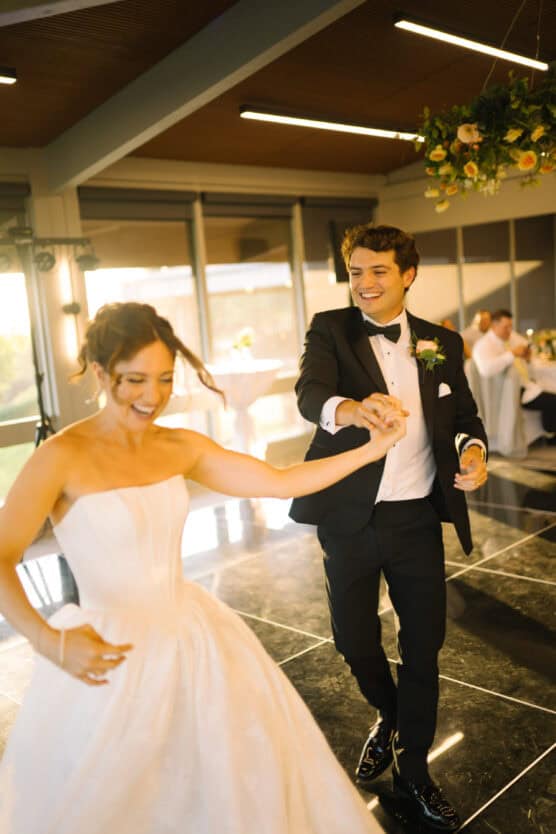 A bride in a white dress and a groom in a black tuxedo joyfully dance together at their wedding reception, smiling and holding hands on a shiny black floor under warm lighting. by Justin Salem Meyer