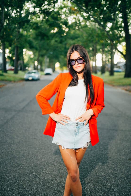 A woman wearing a bright orange blazer, white shirt, and denim shorts stands confidently in the middle of a tree-lined street, with her hands on her hips and sunglasses on. by Justin Salem Meyer