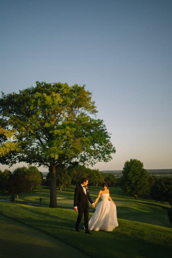 A bride in a white gown and a groom in a black suit walk hand-in-hand on a grassy hill at sunset, with a large tree and a scenic landscape in the background. by Justin Salem Meyer
