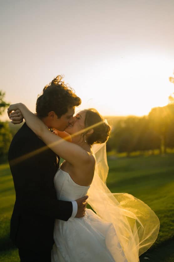 A bride and groom share a romantic kiss outdoors at sunset, with sunlight shining through the bride’s veil and creating a warm, golden glow around them. by Justin Salem Meyer