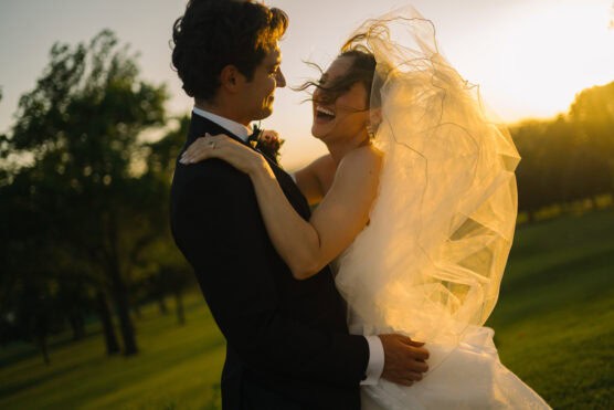 A bride and groom embrace outdoors at sunset, laughing joyfully. The bride’s veil flows in the breeze, illuminated by the golden sunlight, while trees and grass fill the background. by Justin Salem Meyer