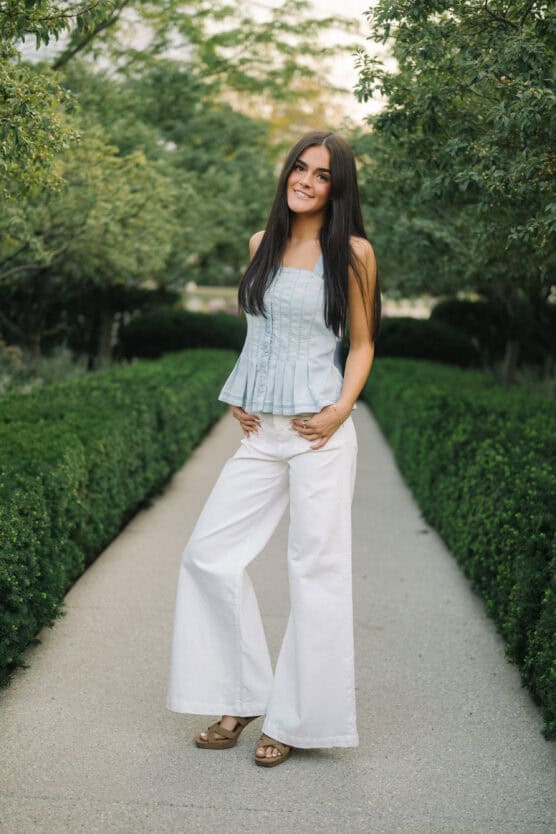 A young woman with long dark hair stands on a garden pathway, wearing a light blue sleeveless top and wide-legged white pants. She smiles at the camera, surrounded by green trees and neatly trimmed hedges. by Justin Salem Meyer