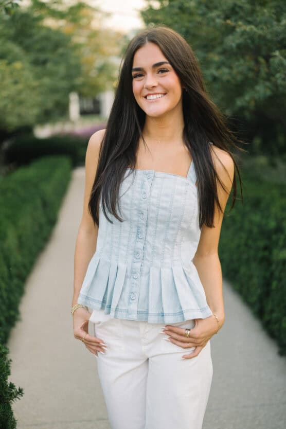 A young woman with long dark hair smiles while standing outdoors on a garden pathway. She is wearing a light blue, sleeveless buttoned top and white pants, with greenery and trees in the background. by Justin Salem Meyer