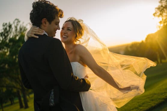 A bride and groom embrace outdoors at sunset, smiling joyfully. The bride’s veil flows in the breeze, and warm sunlight creates a golden, romantic atmosphere around them. Trees and greenery are visible in the background. by Justin Salem Meyer