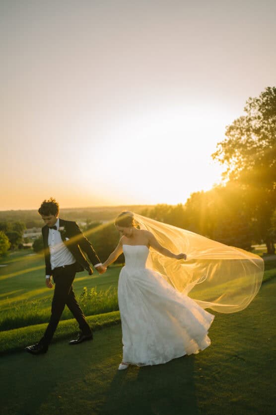 A bride in a white dress and veil walks hand in hand with a groom in a black suit across a grassy field at sunset, with golden sunlight illuminating the scene. by Justin Salem Meyer
