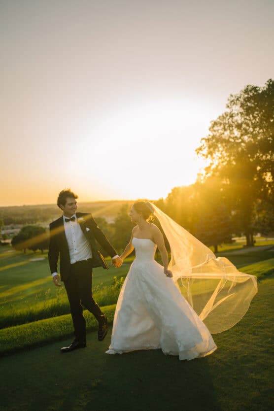 A bride and groom walk hand-in-hand on a grassy field at sunset. The bride wears a strapless white dress with a long veil flowing in the breeze, and the groom is in a black tuxedo. The sun glows warmly behind them. by Justin Salem Meyer