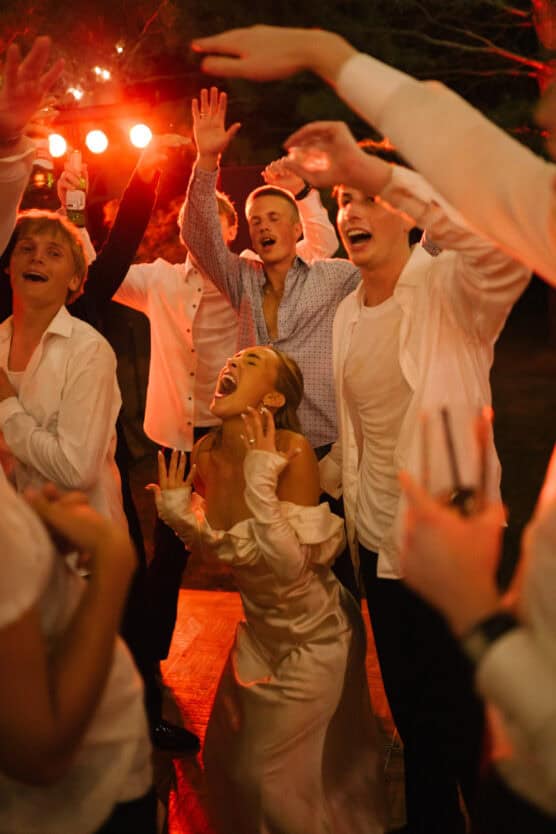 A group of people in white shirts and a woman in a white dress dance and sing enthusiastically under warm, orange lights, with their arms raised and joyful expressions on their faces. by Justin Salem Meyer