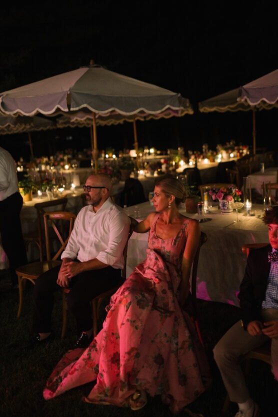 A woman in a pink floral dress sits beside a man in a white shirt at an outdoor evening event with decorated tables, candles, and umbrellas in the background. by Justin Salem Meyer