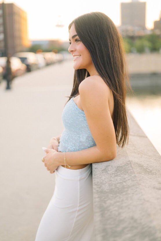 A woman with long dark hair, wearing a light blue sleeveless top and white pants, smiles while leaning on a stone railing outdoors during sunset. Blurred buildings and water are in the background. by Justin Salem Meyer
