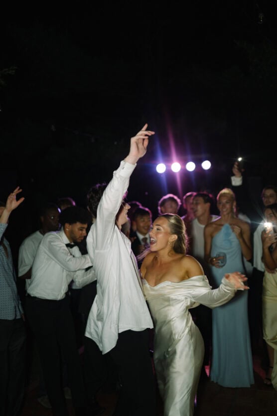 A bride in a white dress dances joyfully with guests at a nighttime celebration, surrounded by people in formal attire, with colorful stage lights in the background. by Justin Salem Meyer