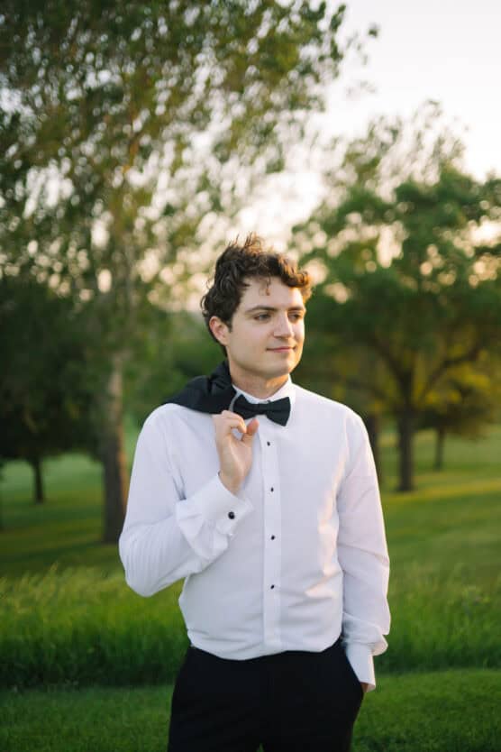 A young man wearing a white dress shirt, black bow tie, and black pants stands outdoors on green grass, holding his jacket over his shoulder. Trees and soft sunlight are in the background. by Justin Salem Meyer