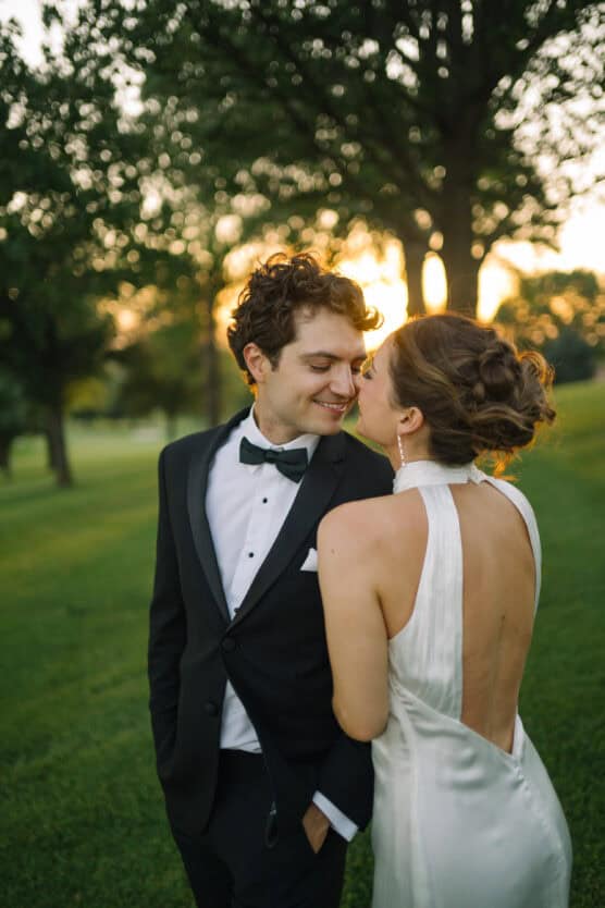A couple dressed in formal wedding attire stands close together outdoors at sunset, smiling and gazing at each other amid green grass and trees. by Justin Salem Meyer