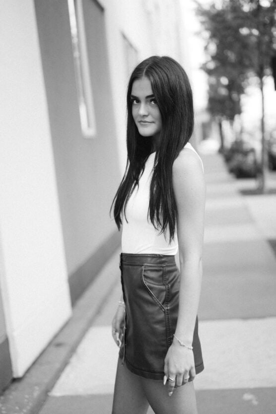 A young woman with long dark hair, wearing a sleeveless white top and a dark skirt, stands on a city sidewalk and looks toward the camera. The photo is in black and white. by Justin Salem Meyer