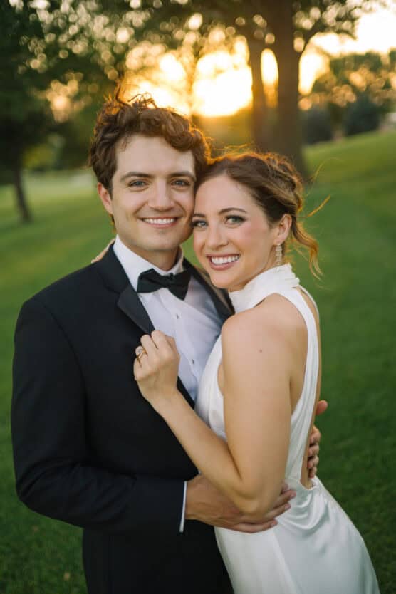 A smiling couple dressed in formal wedding attire embrace outdoors on a grassy lawn at sunset, with trees and soft sunlight in the background. by Justin Salem Meyer