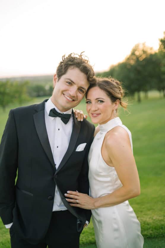 A smiling couple poses outdoors at sunset, the man in a black tuxedo and bow tie, and the woman in a white sleeveless gown with her hand on his chest, standing on green grass with trees in the background. by Justin Salem Meyer