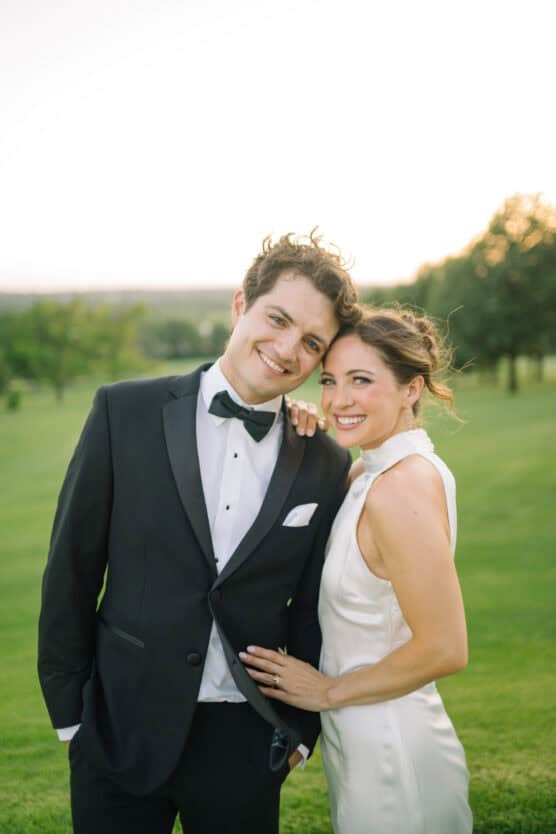 A smiling couple poses outdoors on a grassy field at sunset; the man wears a black tuxedo, and the woman wears a sleeveless white dress, leaning her head on his shoulder. by Justin Salem Meyer
