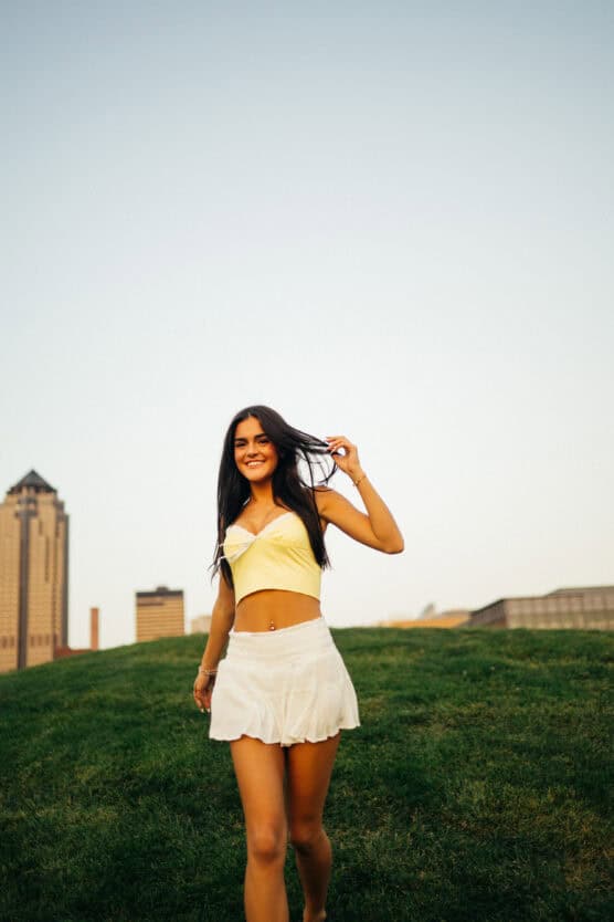A young woman in a yellow crop top and white skirt walks on grass, smiling, with tall buildings and a clear sky in the background. by Justin Salem Meyer
