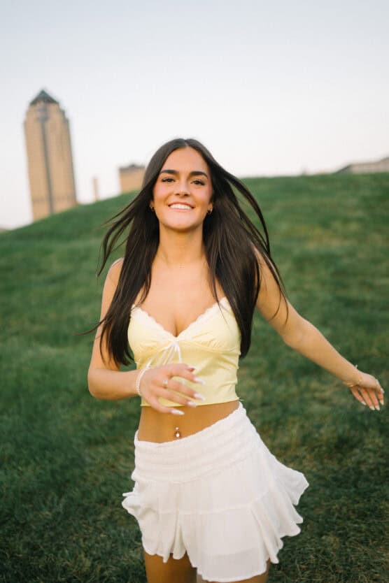 A young woman with long brown hair smiles while standing on green grass, wearing a yellow crop top and a white skirt. A tall building is visible in the blurred background. by Justin Salem Meyer
