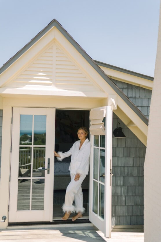 A person in white pajamas with feathered cuffs and hems stands in the doorway of a gray shingled house with a sloped roof, bathed in bright daylight. by Justin Salem Meyer