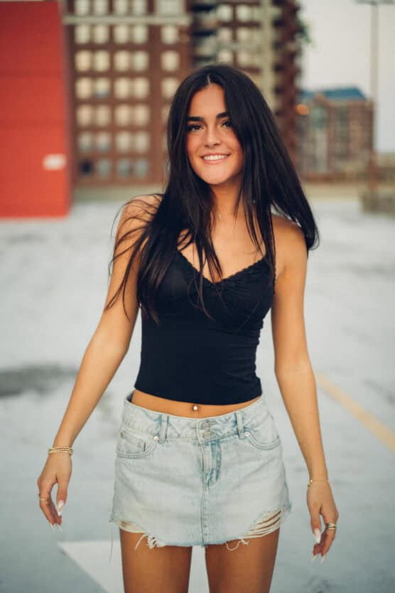 A young woman with long dark hair smiles while standing outdoors. She wears a black camisole top and a light denim skirt with frayed edges. Buildings are blurred in the background. by Justin Salem Meyer