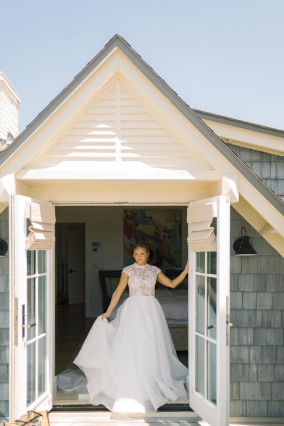 A bride in a white dress stands in the doorway of a house with light gray siding and white trim, framed by an open set of French doors under a gabled roof on a sunny day. by Justin Salem Meyer