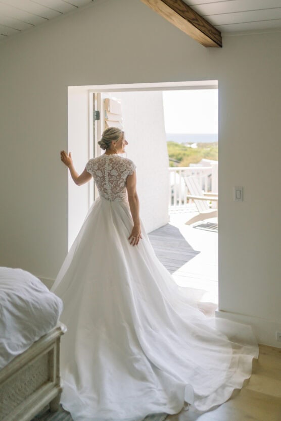 A bride in a white lace wedding gown stands in a sunlit doorway, looking outside onto a balcony. The room is bright and airy with white walls and wooden accents. by Justin Salem Meyer
