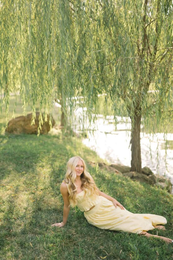 A woman in a pale yellow dress with long blonde hair sits on green grass under a willow tree, with sunlight filtering through the leaves near a pond or riverbank. by Justin Salem Meyer