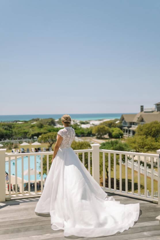 A bride in a white gown stands on a balcony, facing away, overlooking a scenic view of a pool, green landscape, and the ocean under a clear blue sky. by Justin Salem Meyer