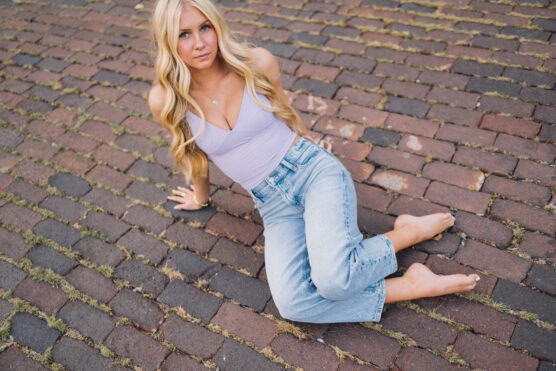 A young woman with long blonde hair sits barefoot on a cobblestone street wearing a light purple top and blue jeans, looking up at the camera with a relaxed expression. by Justin Salem Meyer