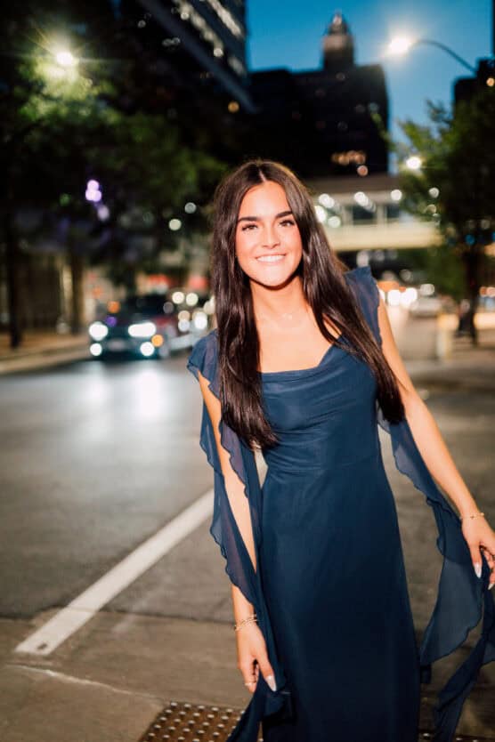 A young woman with long dark hair, wearing a flowing dark blue dress, smiles while standing on a city street at dusk with blurred car lights and buildings in the background. by Justin Salem Meyer