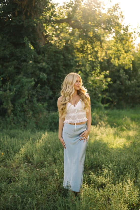 A woman with long blonde hair stands in a sunlit grassy field, wearing a white sleeveless top and a light blue skirt. Trees and greenery surround her, and warm sunlight filters through the leaves. by Justin Salem Meyer