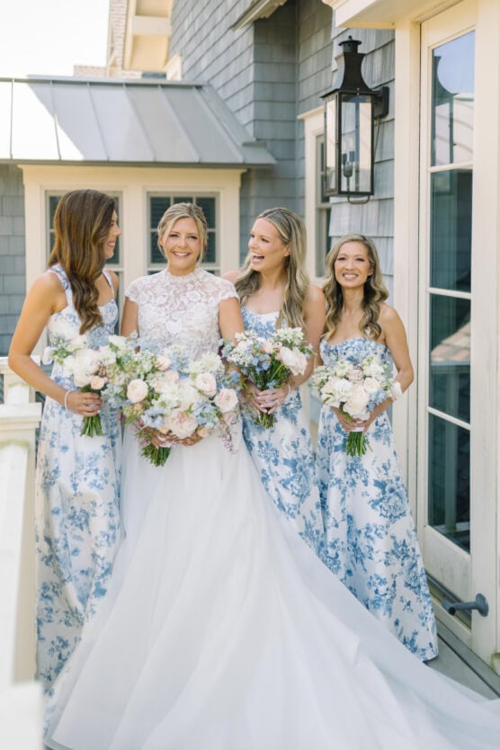 A bride in a white dress stands smiling with three bridesmaids in blue and white floral dresses, all holding bouquets of white and pink flowers on a sunlit porch. by Justin Salem Meyer