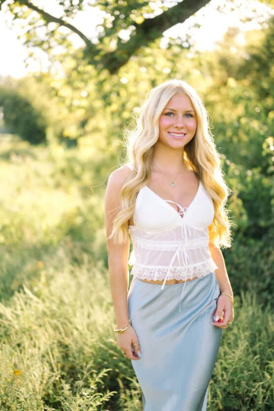 A young woman with long blonde hair wearing a white lace top and a light blue skirt stands outdoors in a sunlit, green, natural setting, smiling at the camera. by Justin Salem Meyer