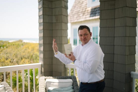 A man in a white shirt stands on a porch, smiling and holding up a small booklet. The background shows greenery, part of a house, and a distant view of the ocean under a clear sky. by Justin Salem Meyer
