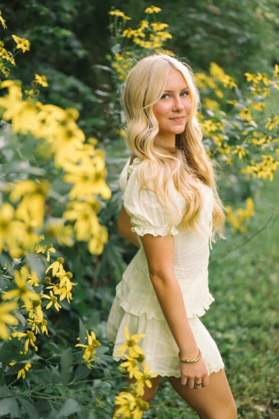 A young woman with long blonde hair in a light-colored dress stands outdoors, surrounded by tall yellow flowers and greenery, smiling softly at the camera. by Justin Salem Meyer